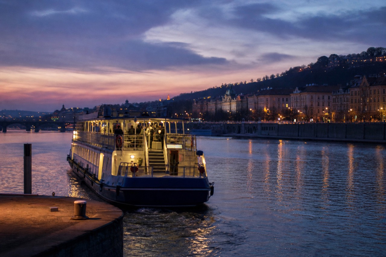 Open-top glass boat on the Vltava at night passing the floodlit Charles Bridge with Prague Castle lit on the hill behind