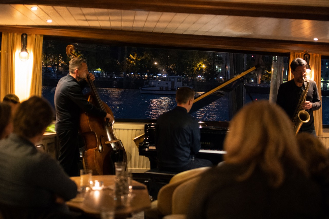 A jazz trio playing on the indoor stage of a double-deck boat on the Vltava at night, diners at tables, the floodlit Prague skyline through the windows