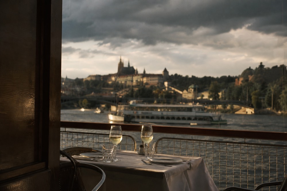 A table set for two on an open boat deck at dusk, wine glasses catching the light, the skyline blurred behind